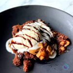 slow cooker chocolate peanut butter brownie on a black plate topped with vanilla ice cream and drizzled with chocolate fudge