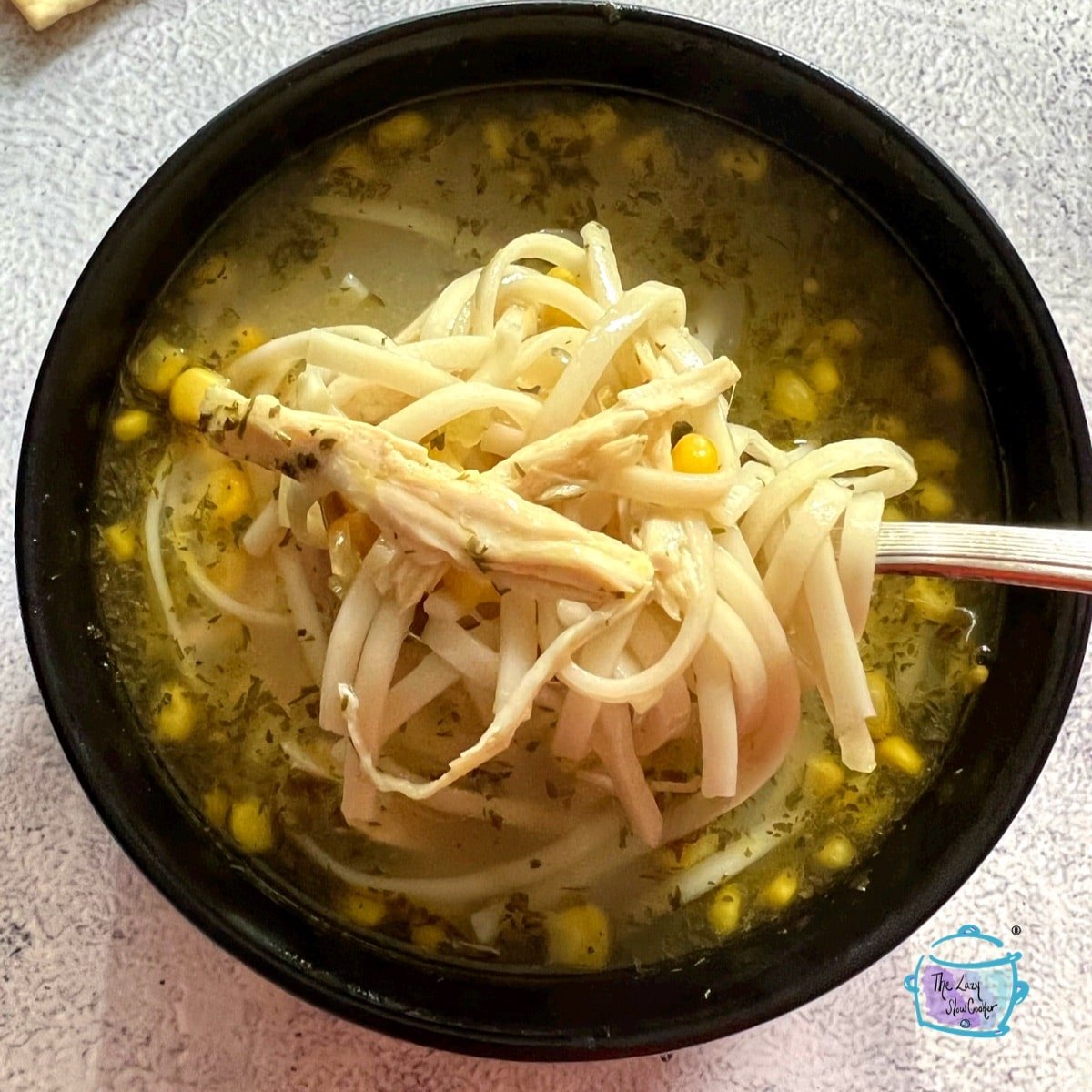 looking down on a spoonful of noodles held over a bowl of chicken soup with corn