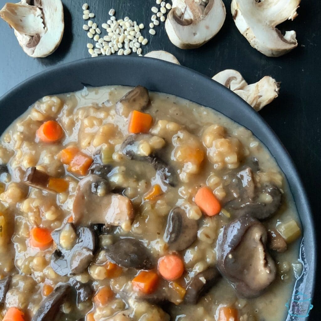 A round black bowl of mushroom barley soup with some raw mushrooms sliced and barley grains laying around it