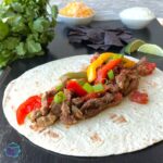 a fajita that is being built with strips of beef and peppers on a flour tortilla