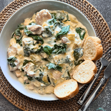 a bowl filled with creamy slow cooked chicken with spinach and artichoke pieces and sliced bread rounds on the side