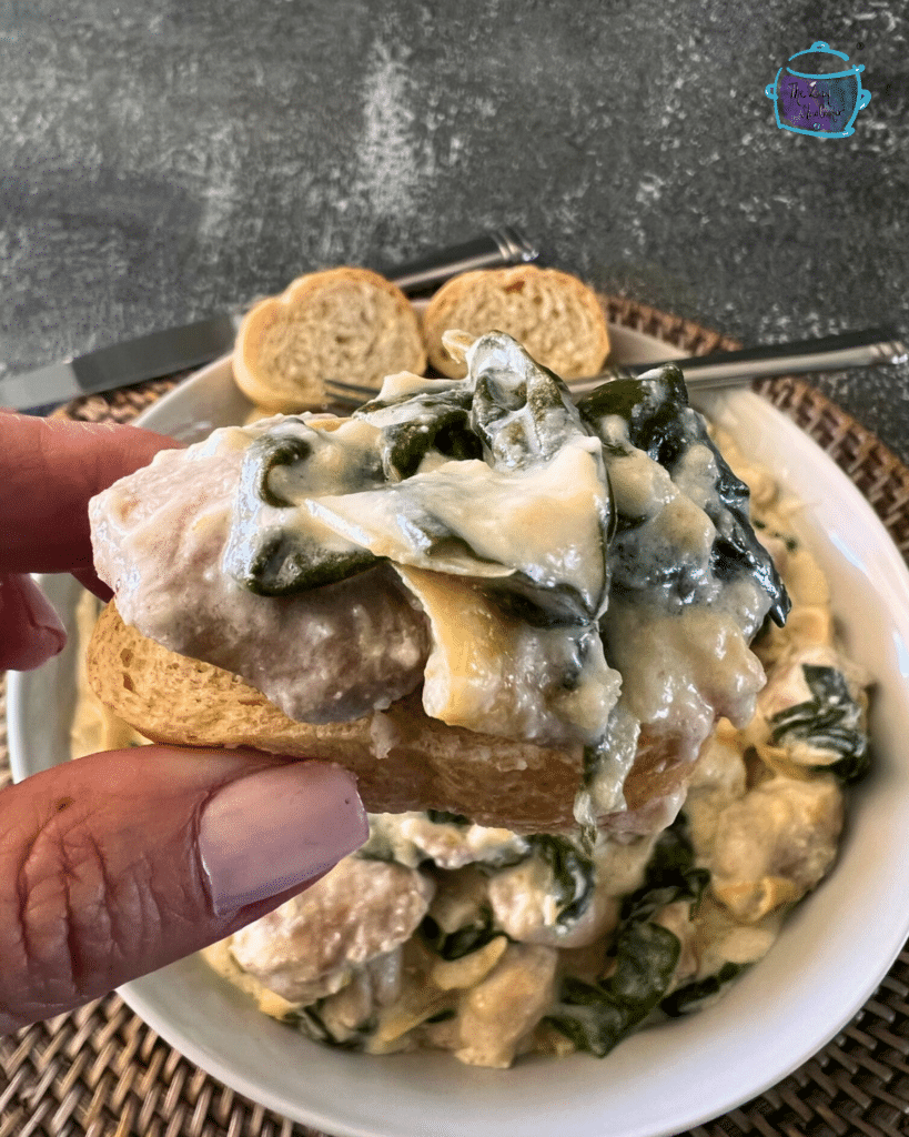 a bowl filled with creamy slow cooked chicken with spinach and artichoke pieces with some on a slice of bread being held up