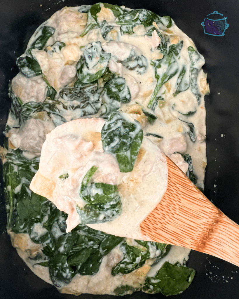 chicken with spinach and artichoke pieces in a crockpot with some held up on a wooden spoon.