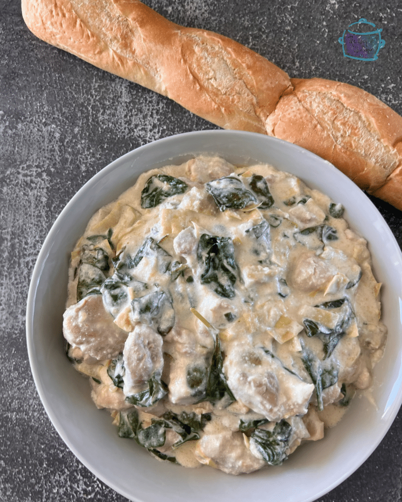 a bowl filled with creamy slow cooked chicken with spinach and artichoke pieces with a long roll beside it,