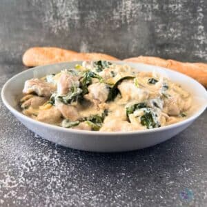 a bowl filled with creamy slow cooked chicken with spinach and artichoke pieces with a long roll beside it,