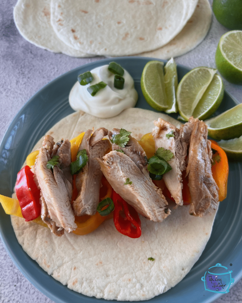 Slow cooker chicken made with tequila and lime on a soft taco shell with veggies, limes wedges and a dollop of sour cream.