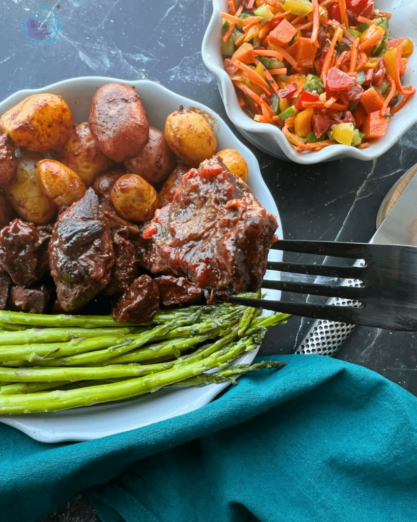 Slow cooker Asian steak bites on a a fork with a plate with a plate of more steak bites, potatoes and asparagus. A salad in on the side