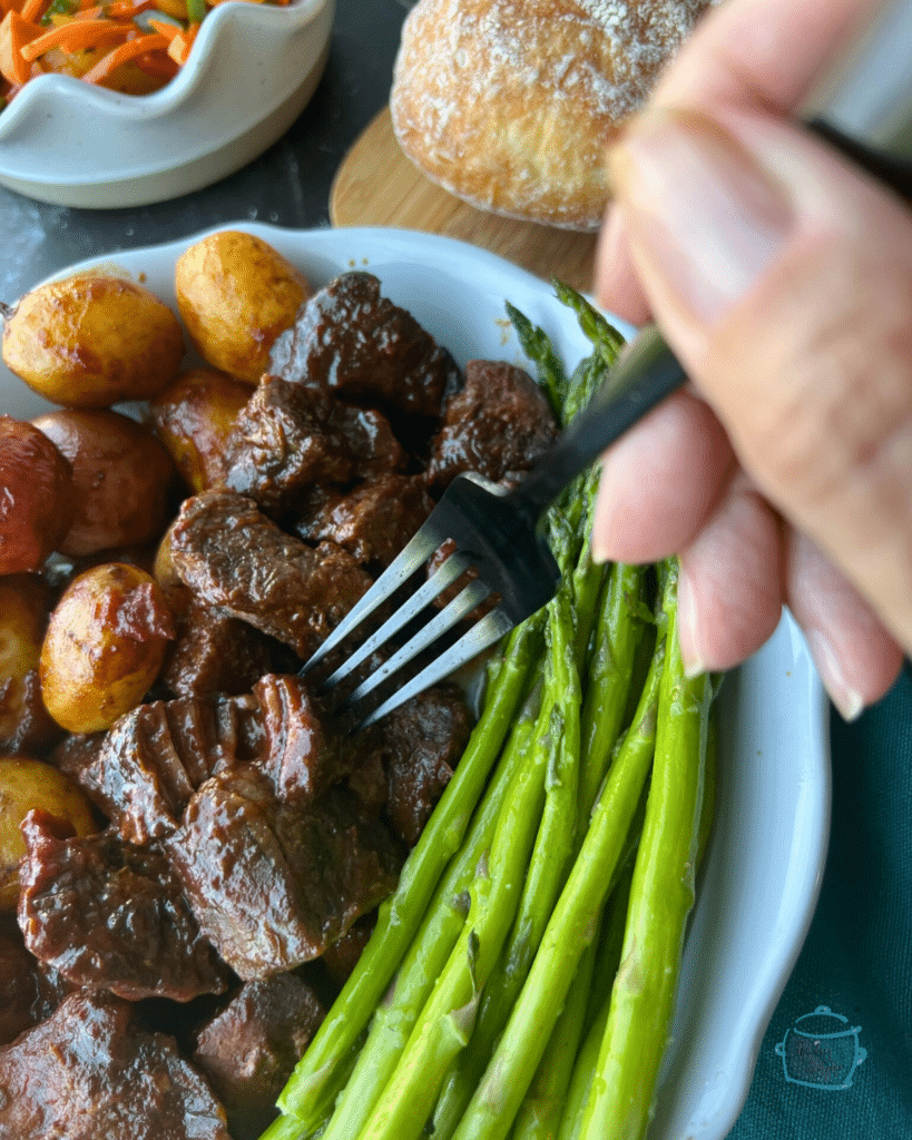 Slow cooker Asian steak bites on a a fork with a plate with a plate of more steak bites, potatoes and asparagus. A salad in on the side