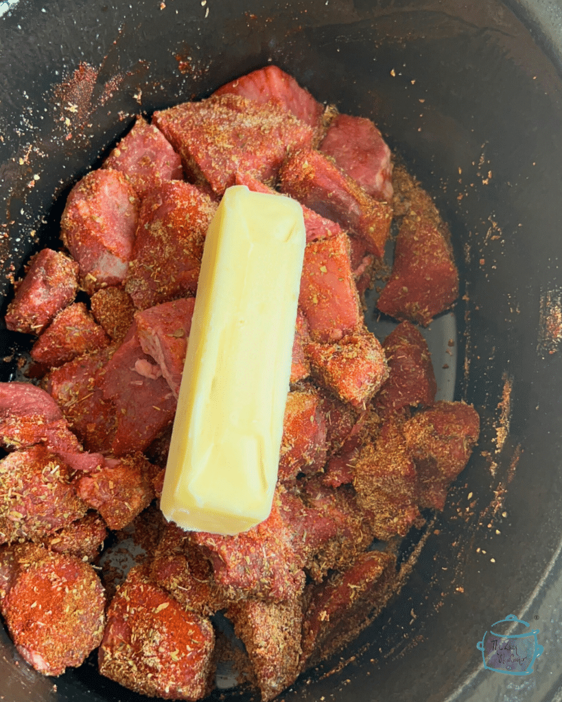 steak bites in crockpot before cooking coated in spices and topped with a stick of butter