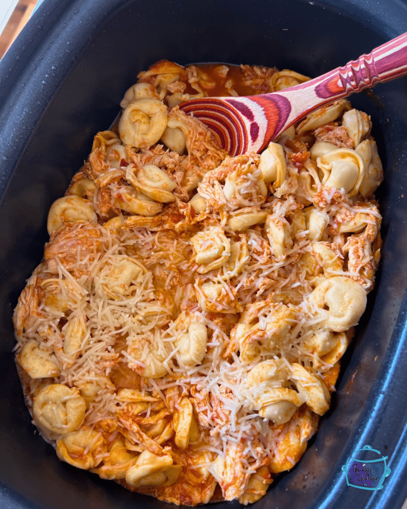 Shredded chicken and tortellini in a slow cooker with a wooden spoon.