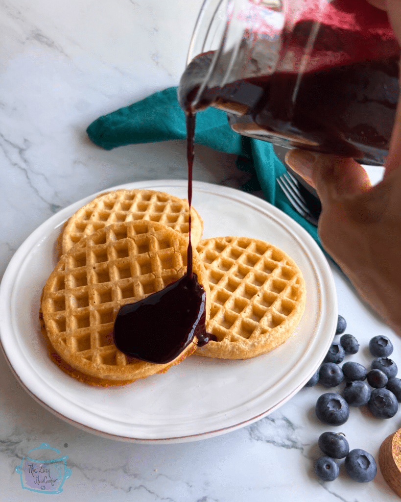 slow cooker blueberry sauce being poured on top of waffles with some fresh blueberries off to the side