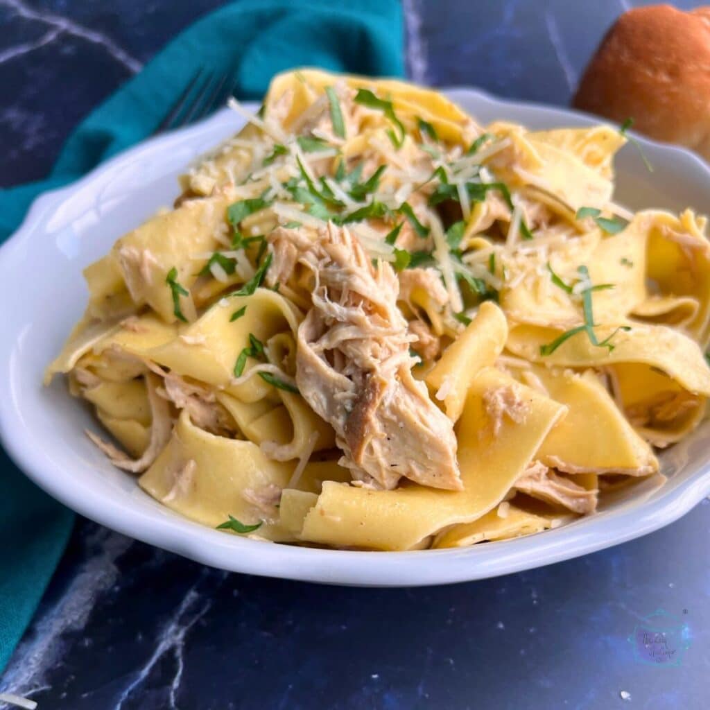 Chicken pieces with wide noodles on a plate topped with chopped fresh herbs and grated parmesan cheese. 