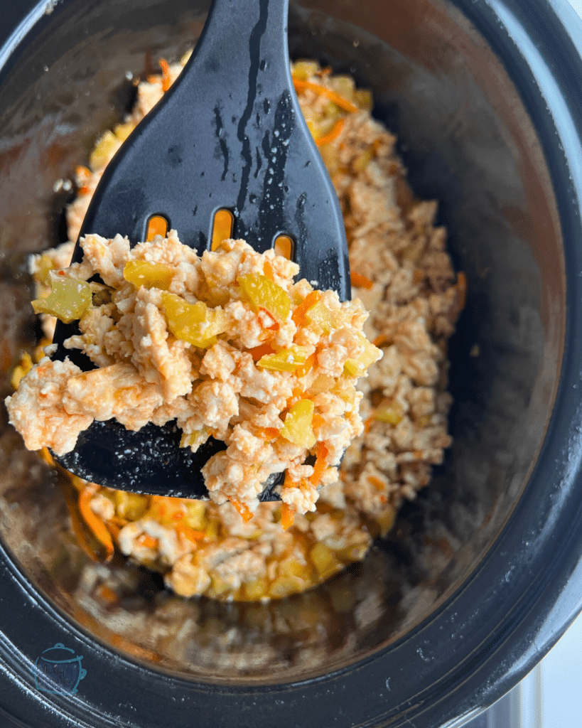 a slow cooker with a spatula in front of it holding some ground cooked ground turkey and veggies in sauce.