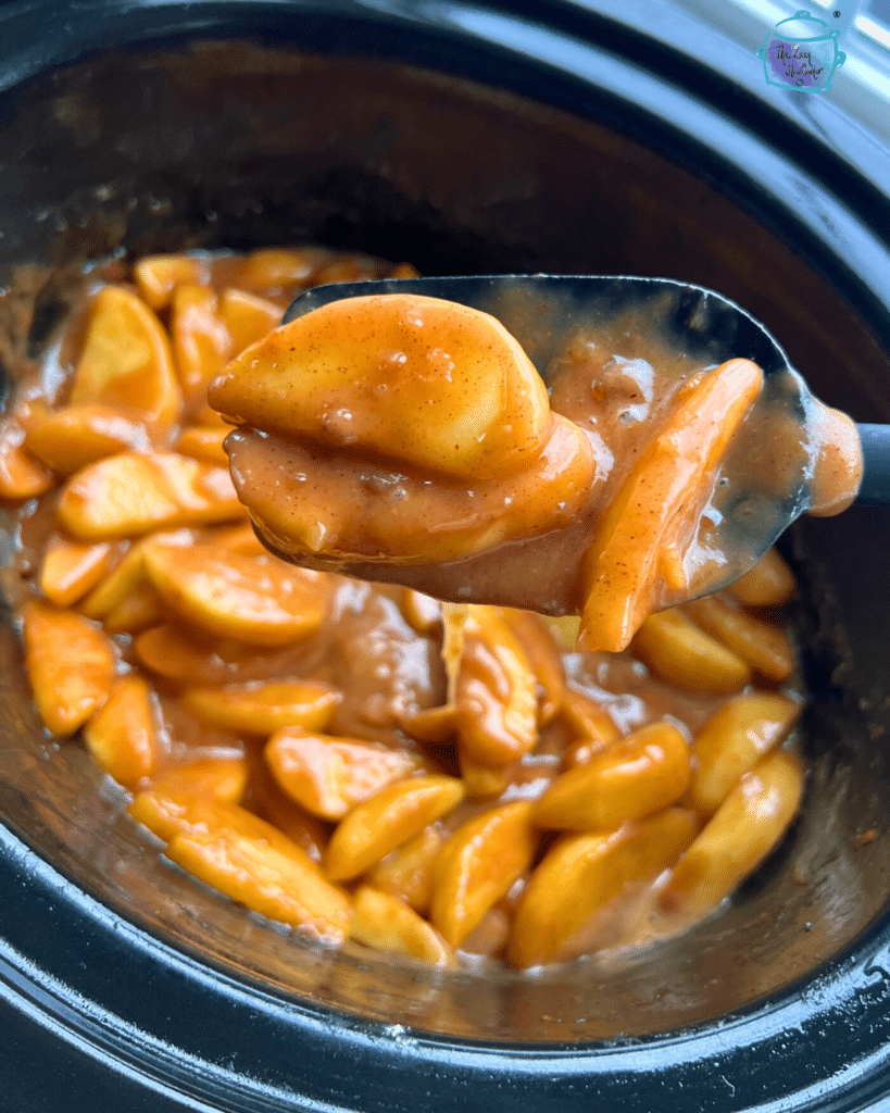 apple pie filling in caramel sauce in a slow cooker after cooking. some is held on a spatula