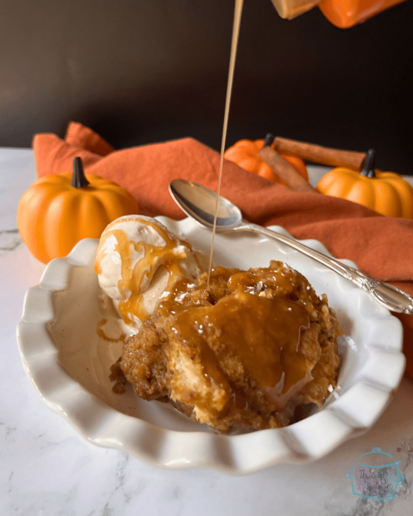 a serving of slow cooker bread pudding in a white bowl with a scoop of vanilla ice cream. With caramel sauce being drizzled on top.