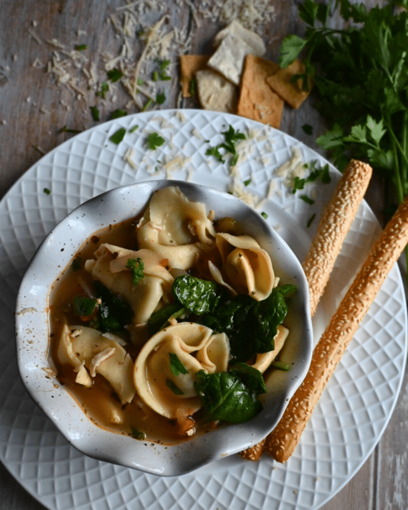 Slow cooker Italian chicken soup with tortellini in a white bowl with bread sticks and fresh parsley on the side.