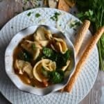 Slow cooker Italian chicken soup with tortellini in a white bowl with bread sticks and fresh parsley on the side.