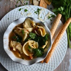 Slow cooker Italian chicken soup with tortellini in a white bowl with bread sticks and fresh parsley on the side.