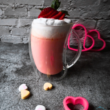 A glass mug filled with pink hot chocolate topped with whipped cream and a sliced up strawberry