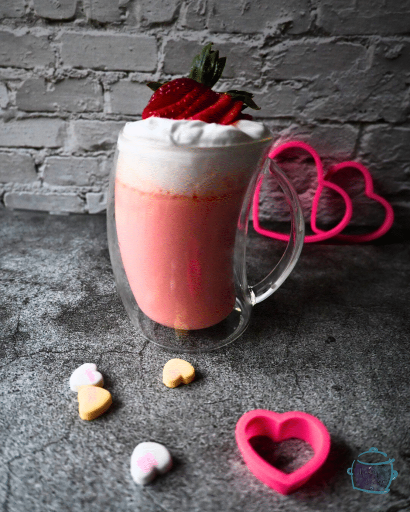 A glass mug filled with pink hot chocolate topped with whipped cream and a sliced up strawberry