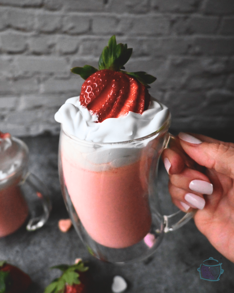 A glass mug filled with pink hot chocolate topped with whipped cream and a sliced up strawberry held up in the air