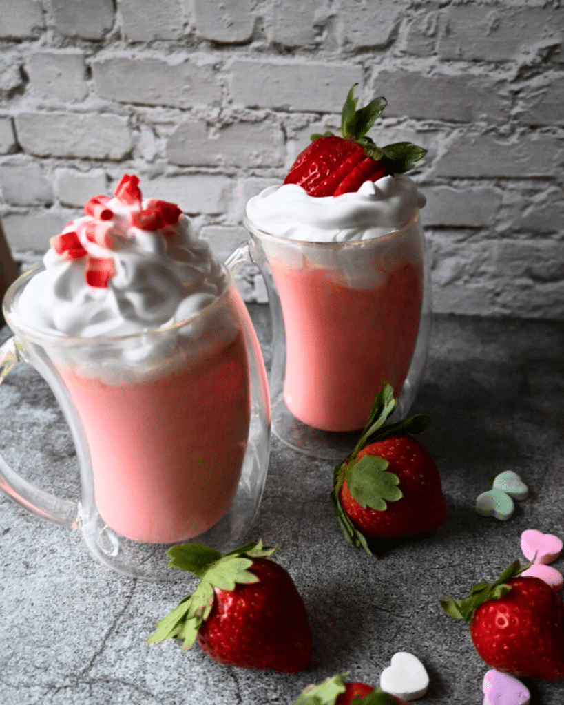 two mugs of crockpot strawberry hot chocolate topped with whipped cream with strawberries lying around