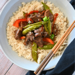 A white plate of teriyaki and sugar snap peas on a bed of rice with chop sticks
