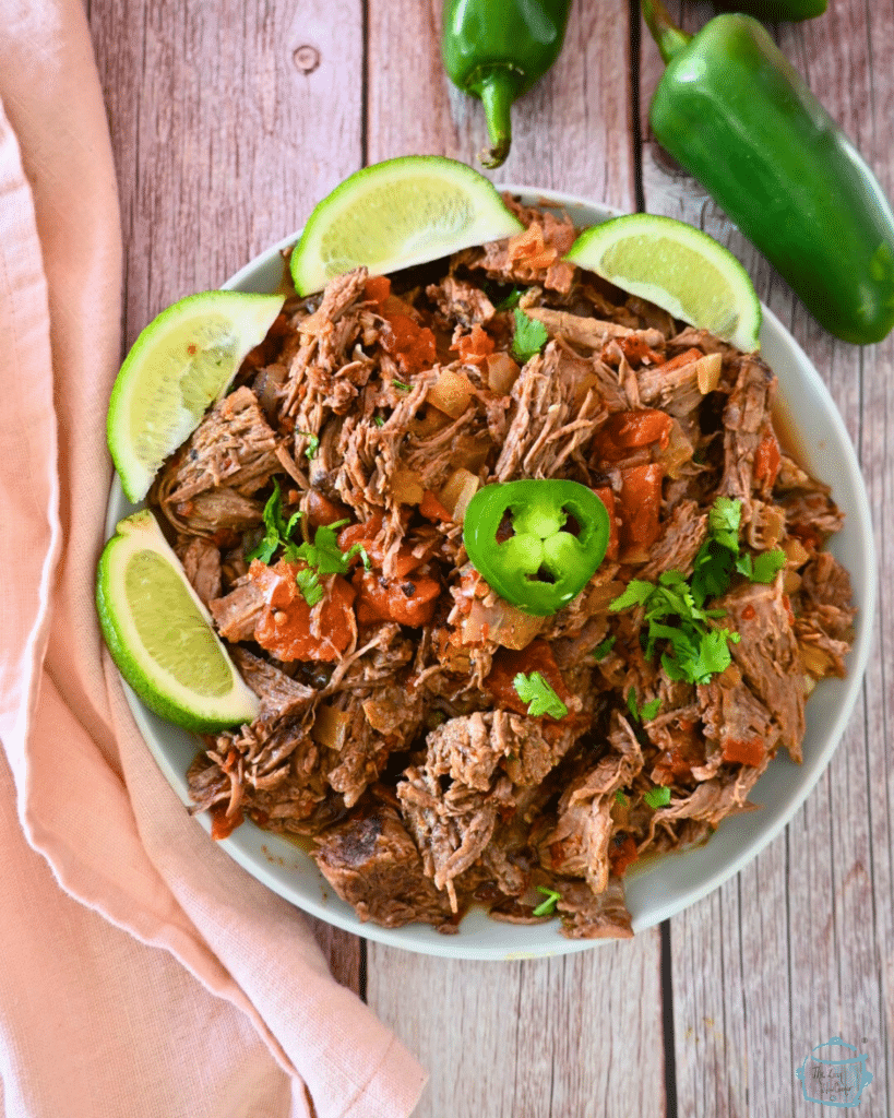 A plate full of slow cooked shredded chipotle beef rimmed with lime wedges and topped with a slice of jalapeño