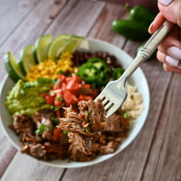 A forkful of shredded chipotle beef held over a plate of chipotle beef with corn beans tomatoes and lime wedges