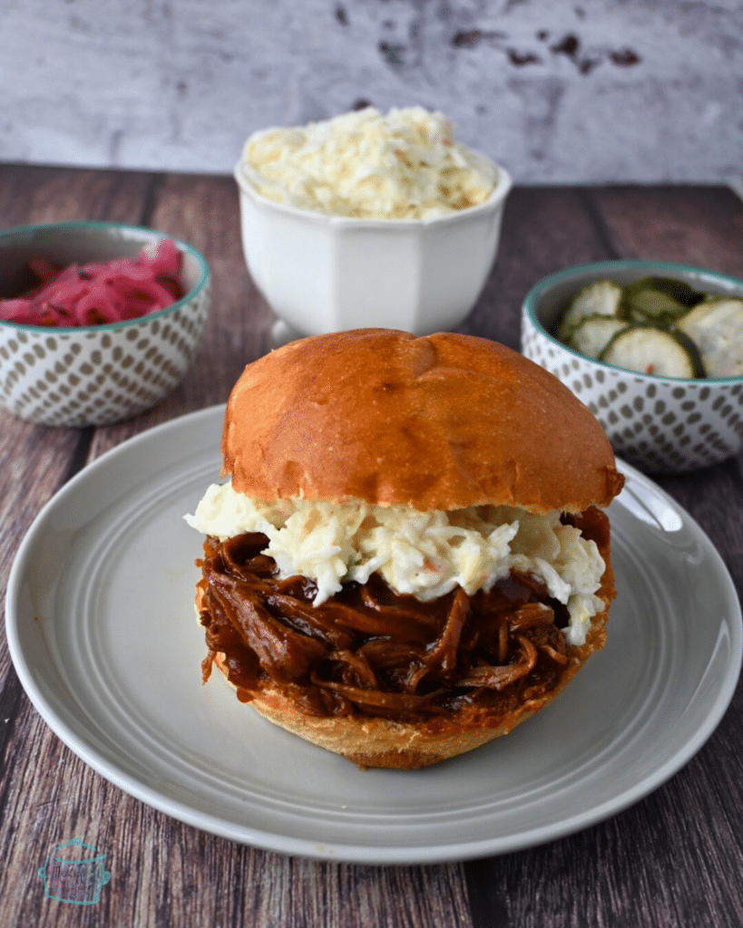 a shredded bbq brisket and coleslaw sandwich with pickles and salad side in the back