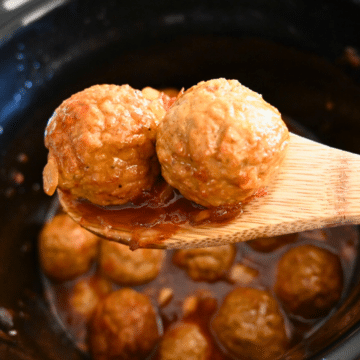 two slow cooked meatballs on a wooden spoon held over a crockpot full of the same