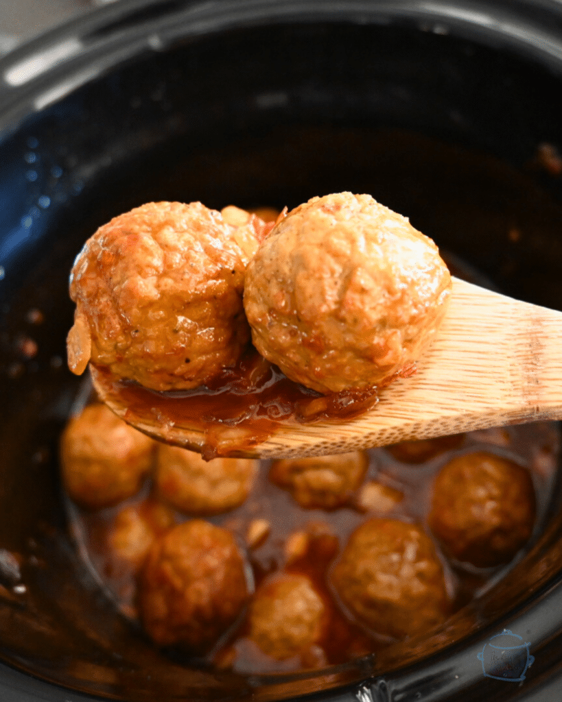 two slow cooked meatballs on a wooden spoon held over a crockpot full of the same