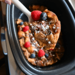 Eggnog bread pudding with berries on a spoon held over a slow cooker full of the same