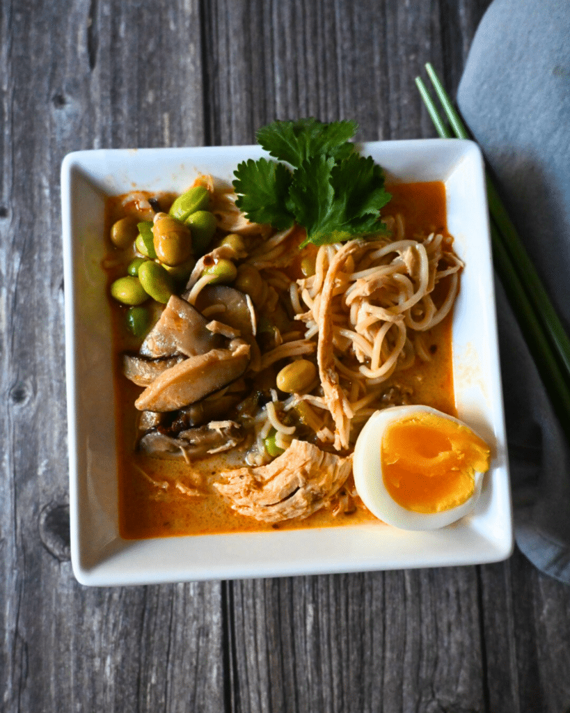 Thai curry ramen with chicken in a square bowl with an egg on the side