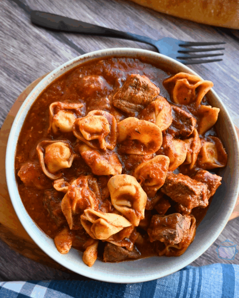 A bowl of slow cooker tortellini pasta and beef chunks in a rich tomato sauce sits on a wooden board, with a fork and bread partially visible in the background.