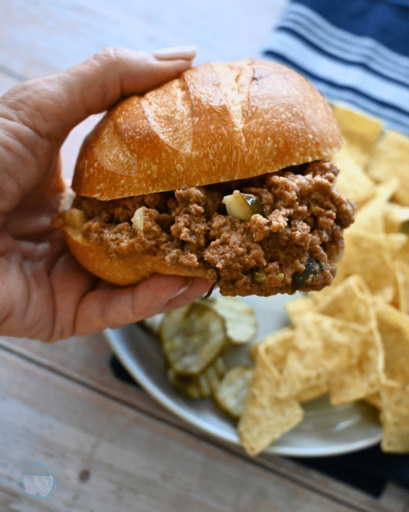 a pickle sloppy joe being held over a plate with chips