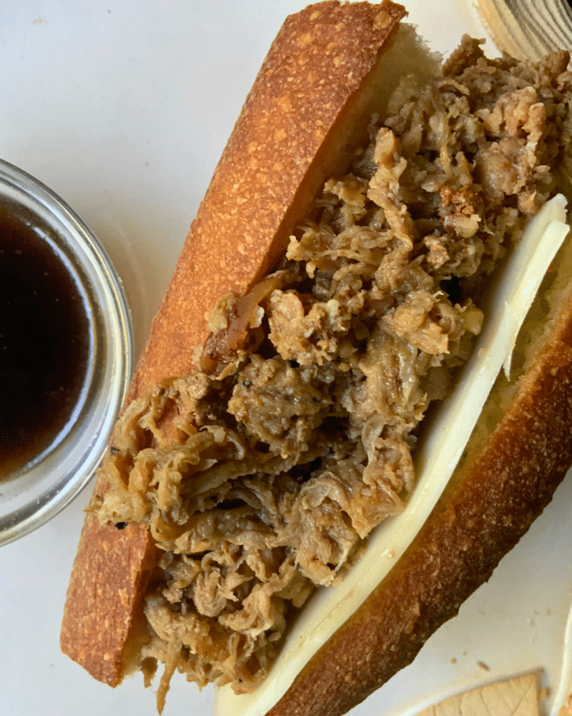 A close-up of a French dip sandwich with shredded beef and melted cheese in a baguette, beside a small bowl of au jus dipping sauce.