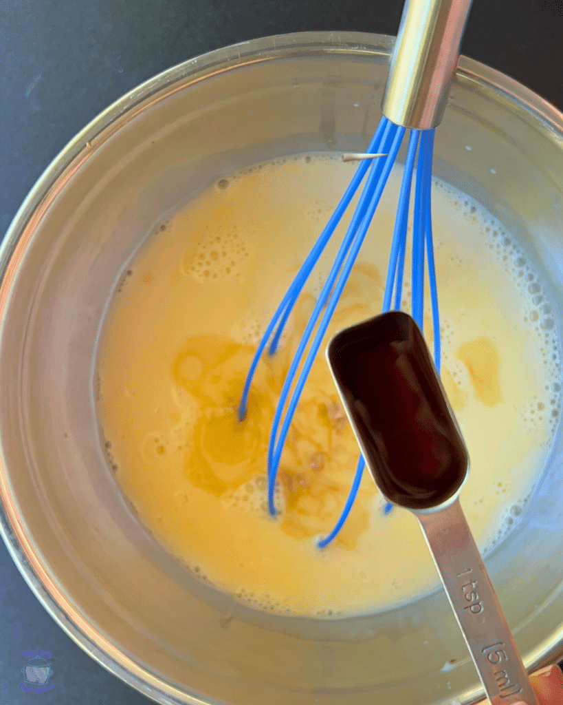 a bowl filled with beaten eggs and milk with a tablespoon of vanilla in front