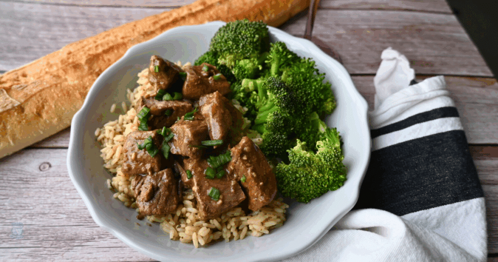 slow cooker steak bites with broccoli on a bed a rice next to a long roll