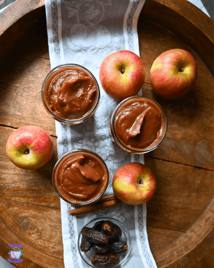 Looking down on three jars of apple butter are arranged on a cloth with fresh apples and cinnamon sticks on a wooden surface.