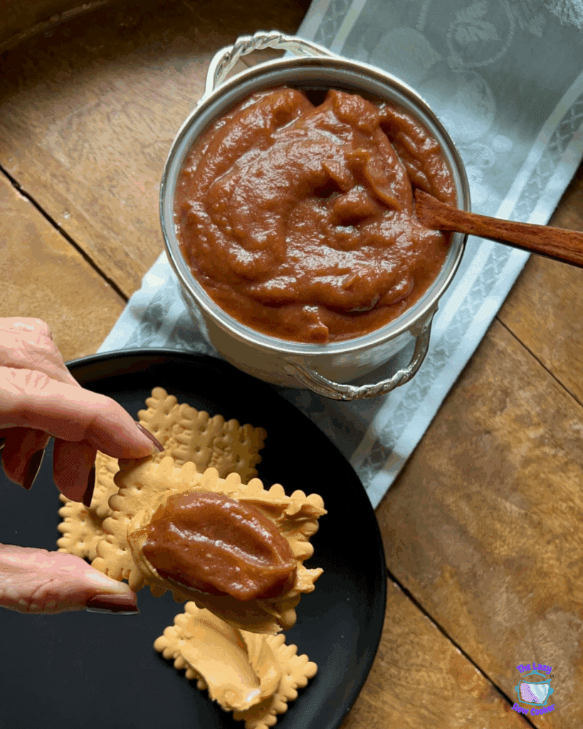 A hand spreads a thick layer of brown apple butter onto a rectangular biscuit, with more biscuits on a black plate and a bowl of apple butter with a wooden spoon nearby.