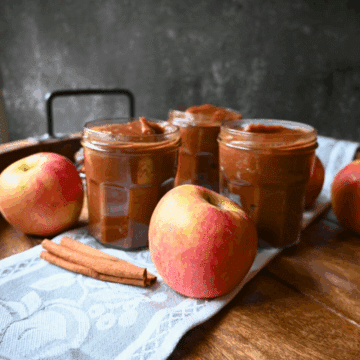 Three jars of apple butter are arranged on a cloth with fresh apples and cinnamon sticks on a wooden surface.