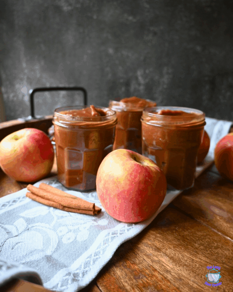 Three jars of apple butter are arranged on a cloth with fresh apples and cinnamon sticks on a wooden surface.