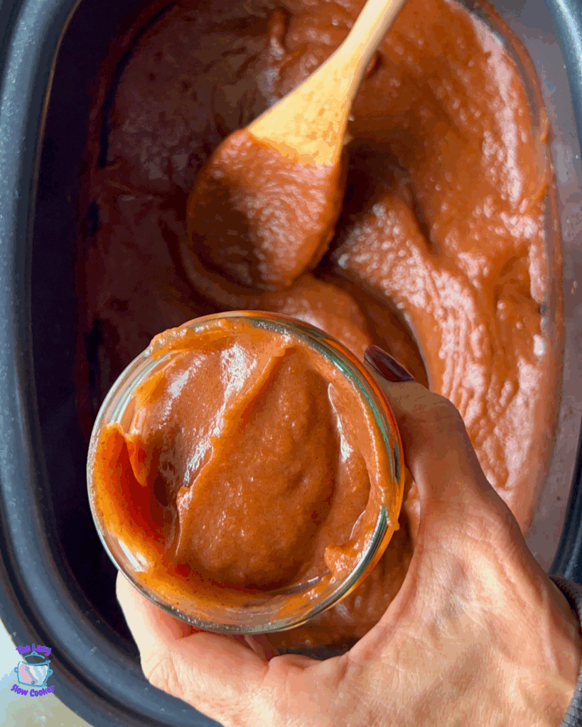 A hand holds a small glass jar filled with thick, brown apple butter. In the background, a wooden spoon rests in a slow cooker containing more apple butter.