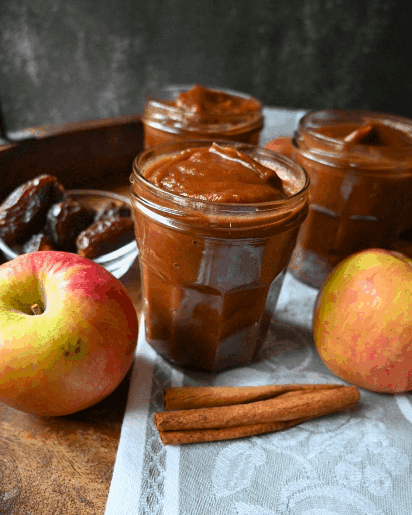 Three jars of apple butter are arranged on a cloth with fresh apples and cinnamon sticks on a wooden surface.