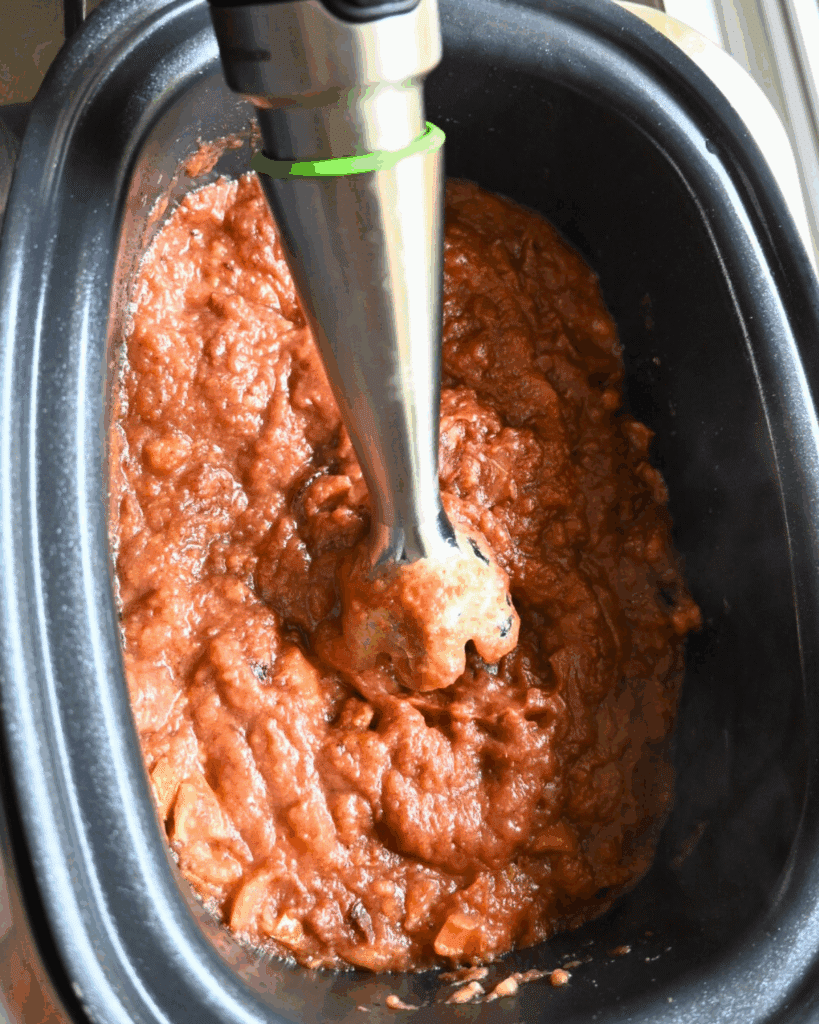 An immersion blender is blending thick, chunky sauce inside a black slow cooker or crockpot.
