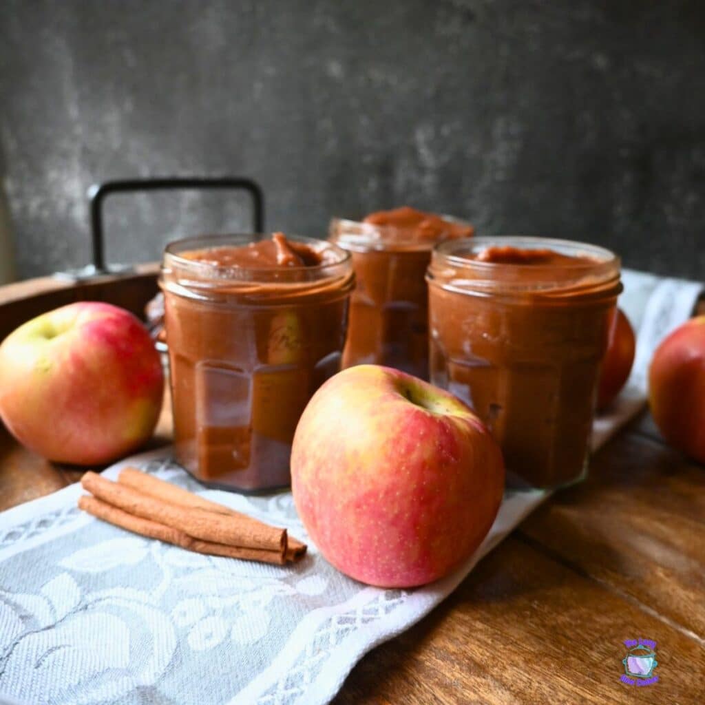 Three jars of apple butter arranged on a cloth with fresh apples and cinnamon sticks on a wooden surface.