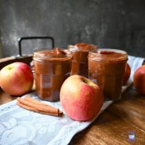 Three jars of apple butter arranged on a cloth with fresh apples and cinnamon sticks on a wooden surface.