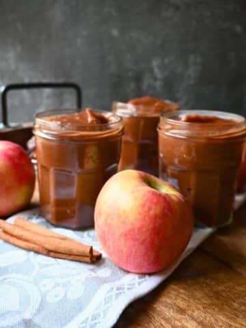 Three jars of apple butter arranged on a cloth with fresh apples and cinnamon sticks on a wooden surface.