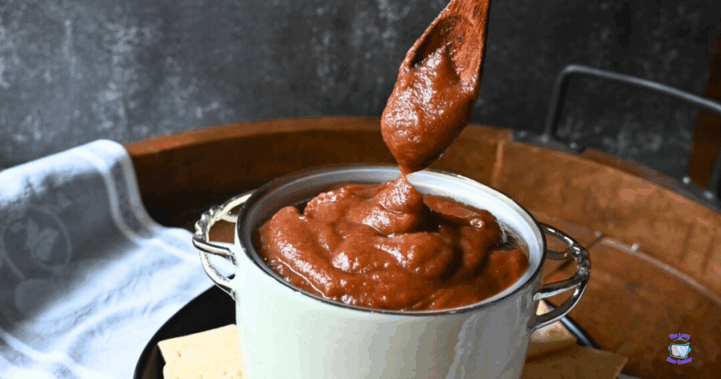 A thick, brown spread is being scooped with a wooden spoon from a white pot with metal handles, set on a wooden tray with a white cloth in the background.